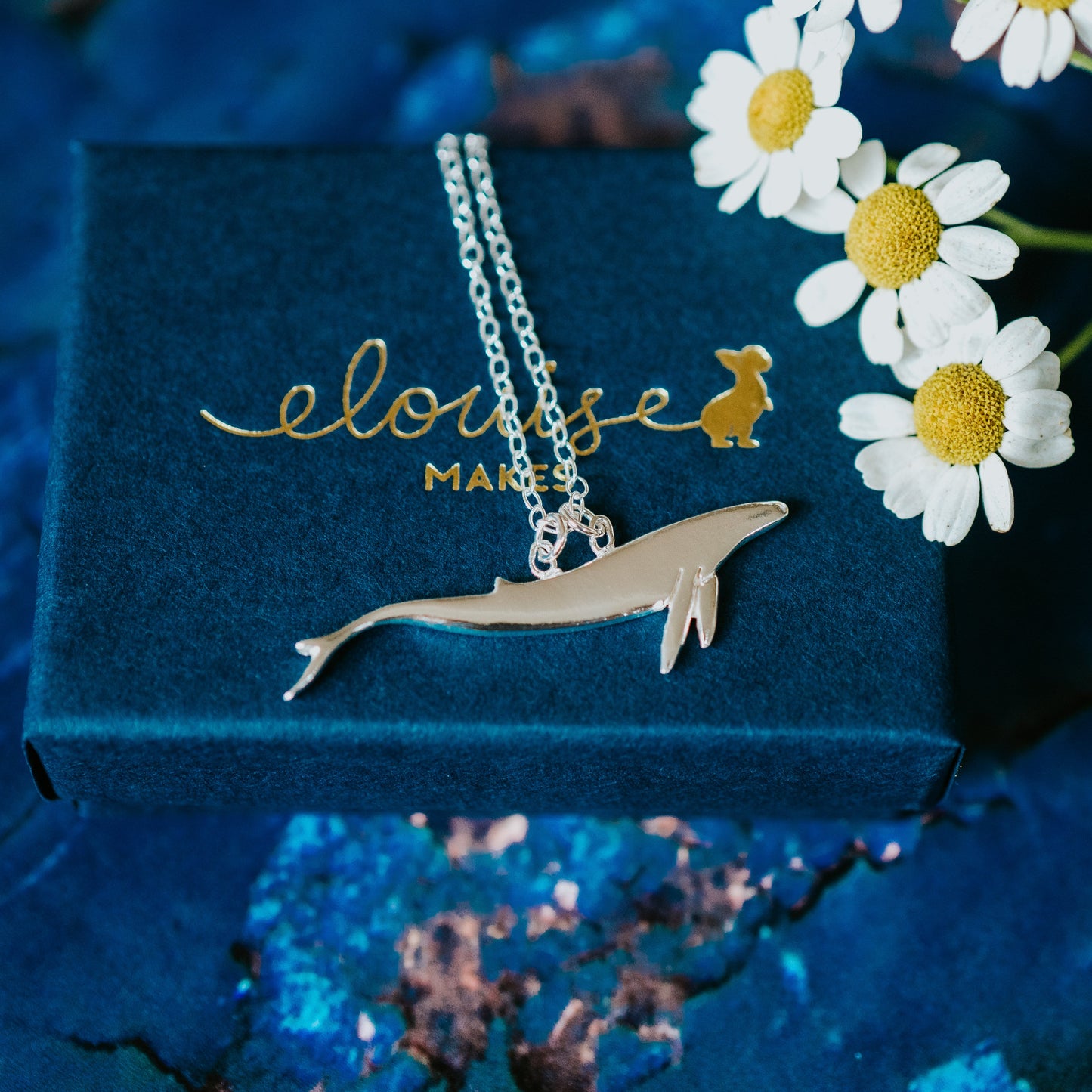Silver necklace with a dolphin pendant on a blue box with 'eloise' branding, surrounded by white flowers.